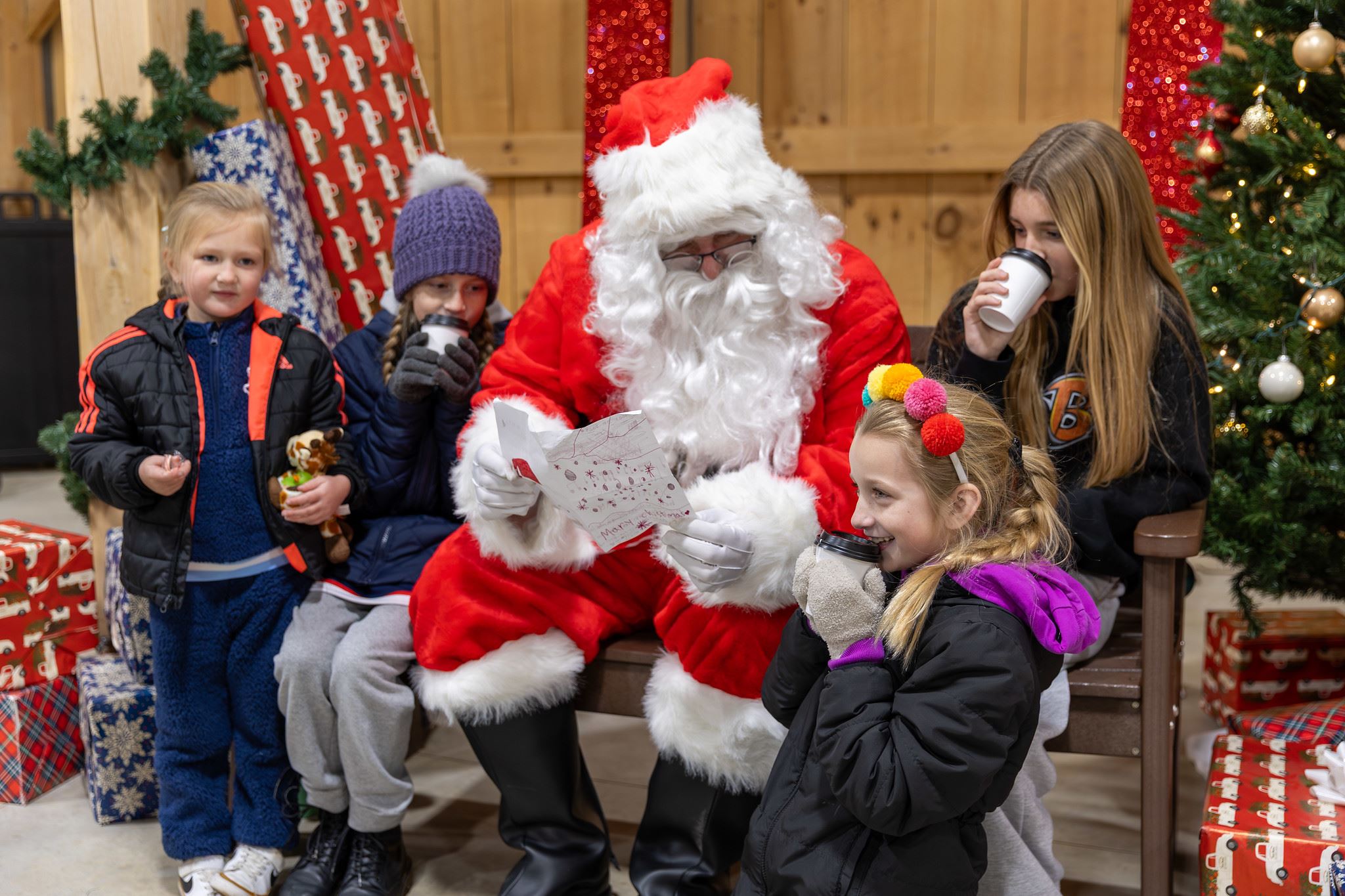 Children gather around Santa as he reads a letter while they sip hot chocolate and smile happily.