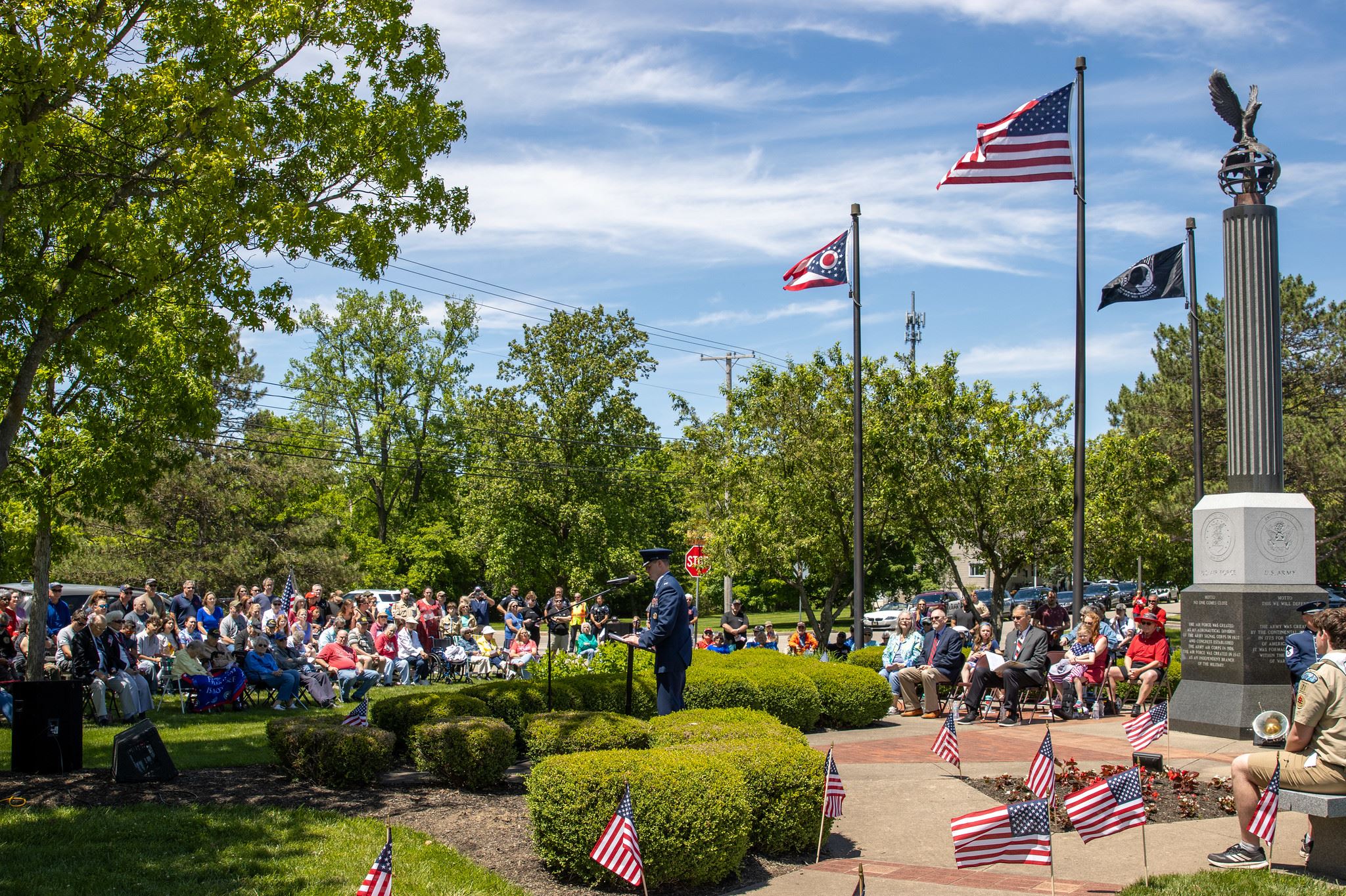 A large crowd listens to a speaker at an outdoor Memorial Day ceremony with flags flying overhead.