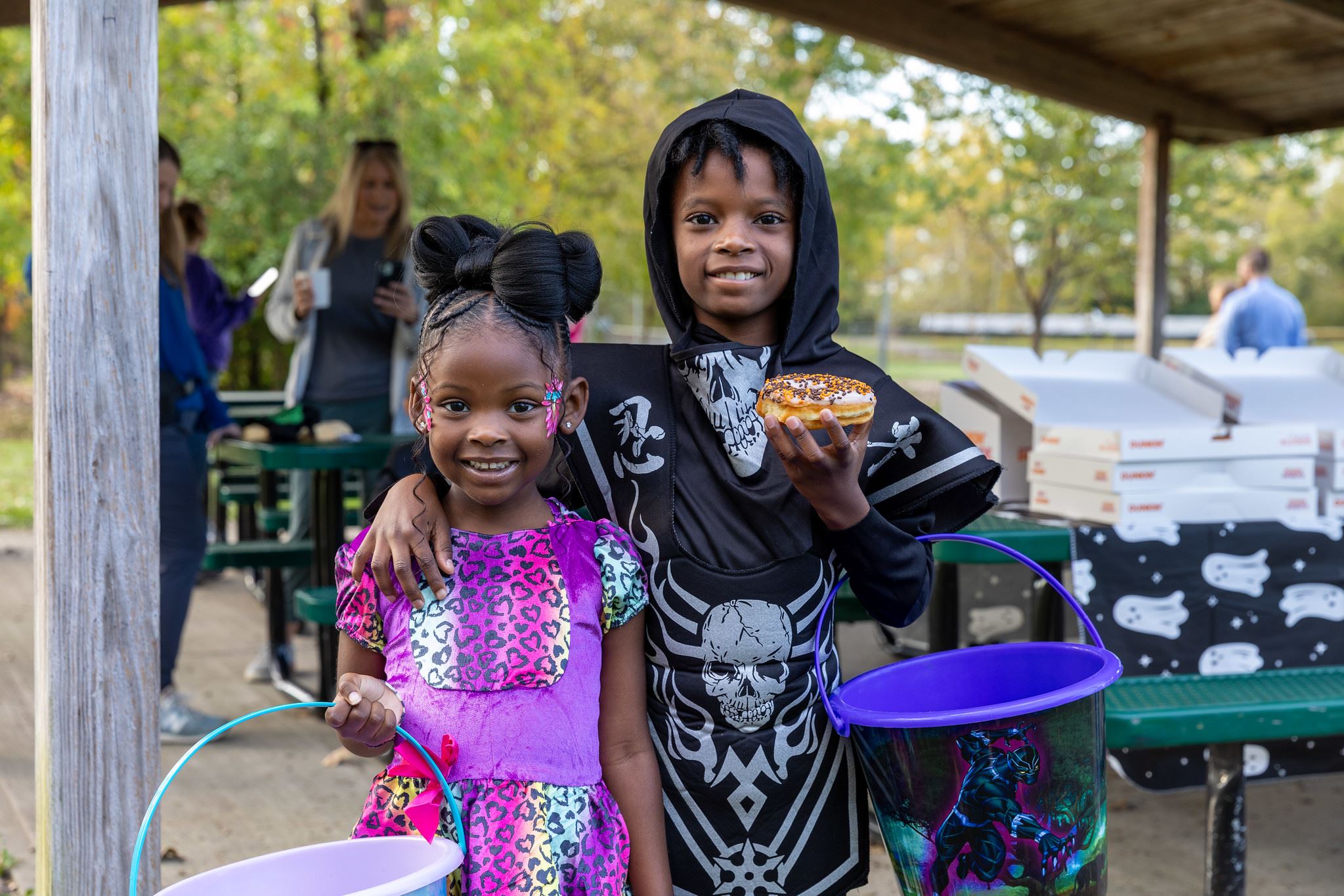Two smiling children in colorful costumes pose with treat buckets, one holding a festive donut.